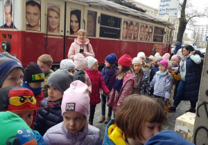 Grupa dzieci stoi na chodniku. Za nimi stoi czerwony tramwaj z afiszami teatralnymi